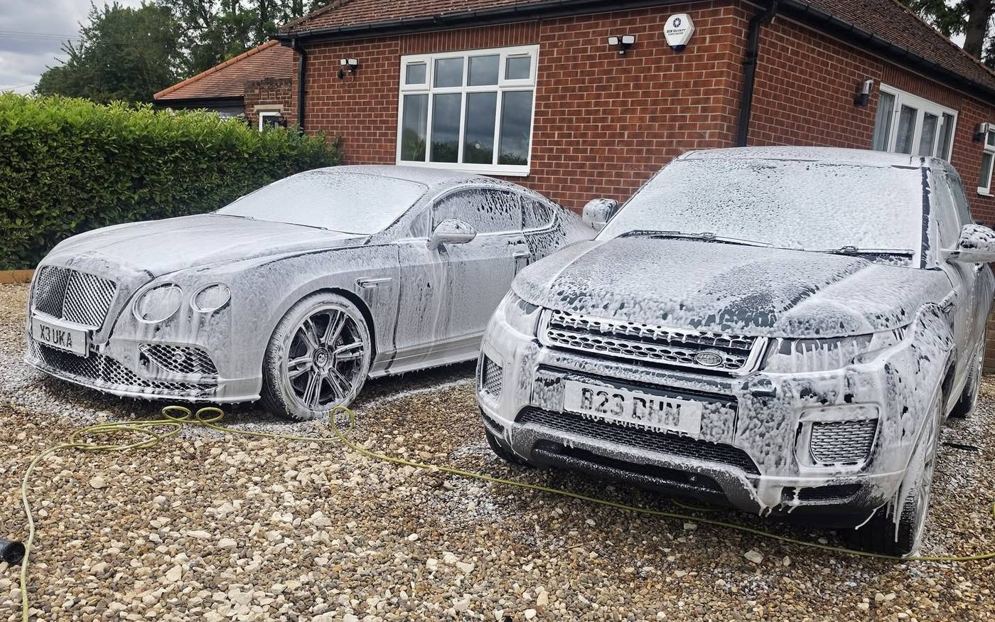 A Bentley and a Range Rover Evoque covered in thick snow foam on a driveway, mid-wash