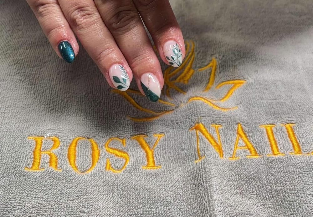 A hand with teal leaf-pattern nail art resting on the salon's own embroidered towel, reading ROSY NAILS in gold thread
